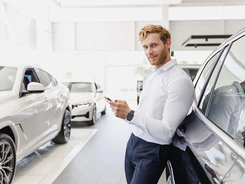 Mann mit Smartphone lehnt an einem Auto: Unfallwagen erkennen vor dem Kauf