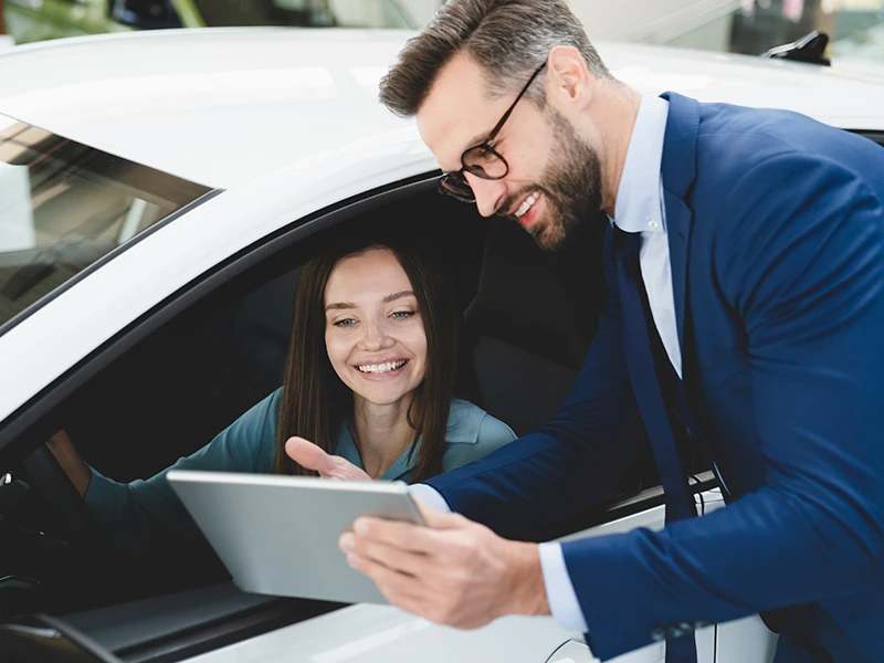 Verkäufer zeigt einer Frau im Auto ein Tablet: Fahrzeughistorie per Fahrgestellnummer digital prüfen