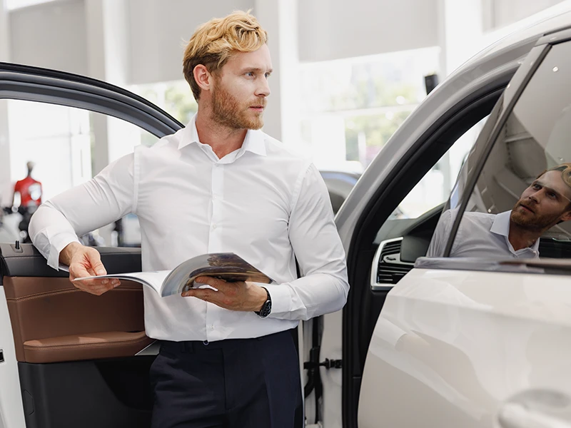 Mann steht mit Dokument an geöffneter Fahrertür: Fahrzeughistorie checken vor dem Kauf