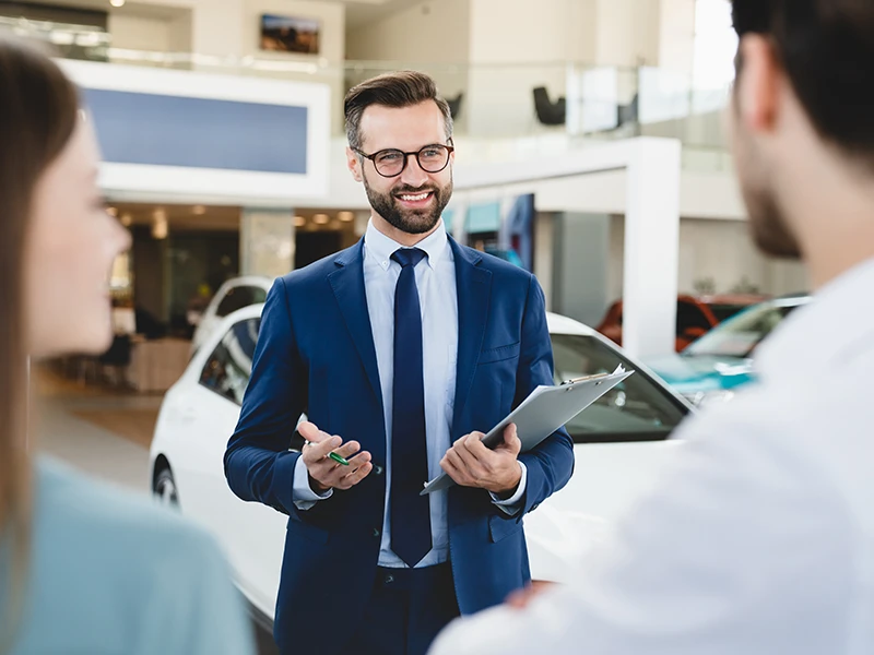 Verkäufer spricht mit einem Paar auf einem Gebrauchtwagenhof: Kilometerstand prüfen und Tachomanipulation erkennen